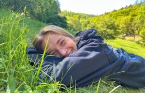 Unmasking: Zu sehen ist ein aktuelles Foto der Autorin. Sie liegt auf dem Bauch auf einer Wiese, fühlt sich wohl und lächelt in die Kamera