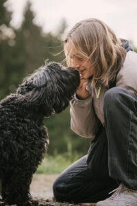 Erfahrungsbericht Autismus Diagnose im Erwachsenenalter: Zu sehen ein aktuelles Foto der Autorin mit ihrem Hund. Der Hund ist schwarz mit langem lockigen Haar und gibt ihr einen Kuss auf die Nase. sie kniet auf dem Boden und lächelt.