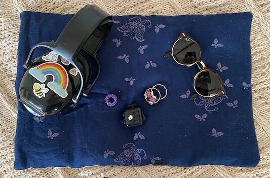 Unmasking: Zu sehen ist ein dunkelblaues gewichtetes Kissen, das mit lila Schmetterlingen bestickt ist. Darauf liegt ein schwarzer Gehörschutz mit bunten Stickern, eine Sonnenbrille und drei Fidget Toys