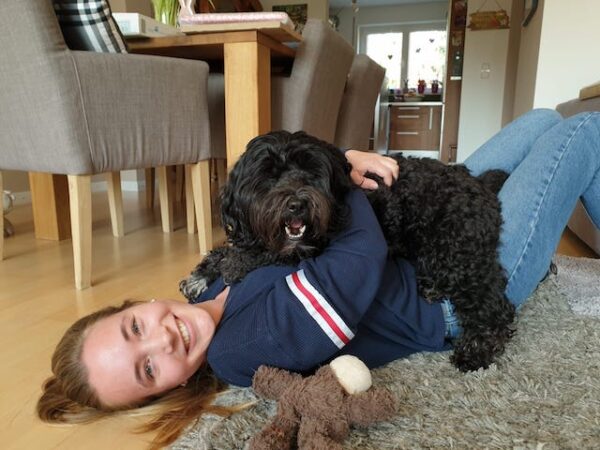 Burnout bei Autismus: Foto von Marlene und ihrem Hund. Das Bild ist im elterlichen Wohnzimmer aufgenommen. Sie liegt auf dem Boden und lächelt in die Kamera, auf ihrem Bauch liegt der Hund und lächelt ebenfalls in die Kamera.