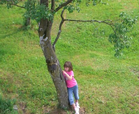 Erfahrungen mit AuDHD: Zu sehen ist ein Foto von Marlene im Alter von 7 Jahren. Sie steht alleine auf einer Wiese an einem Baum angelehnt. Vertieft untersucht sie dabei die Rinde des Baums.