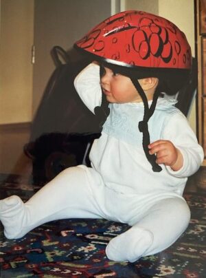 Leben mit AuDHD: Kinderfoto von Marlene. Sie ist ca. 10 Monate alt und sitzt mit einem viel zu großen roten Fahrradhelm mit Ballons auf dem Boden.