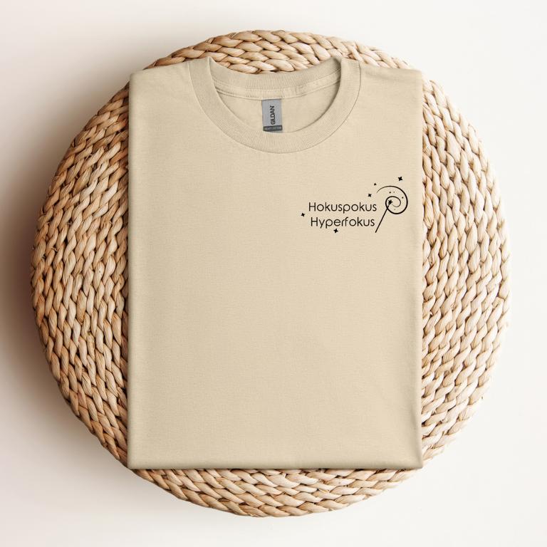 Gefaltetes T-Shirt mit minimalistischer Zauberstab-Grafik und dem Schriftzug „Hokuspokus Hyperfokus“ – humorvolles Design für ADHS & Neurodivergenz.