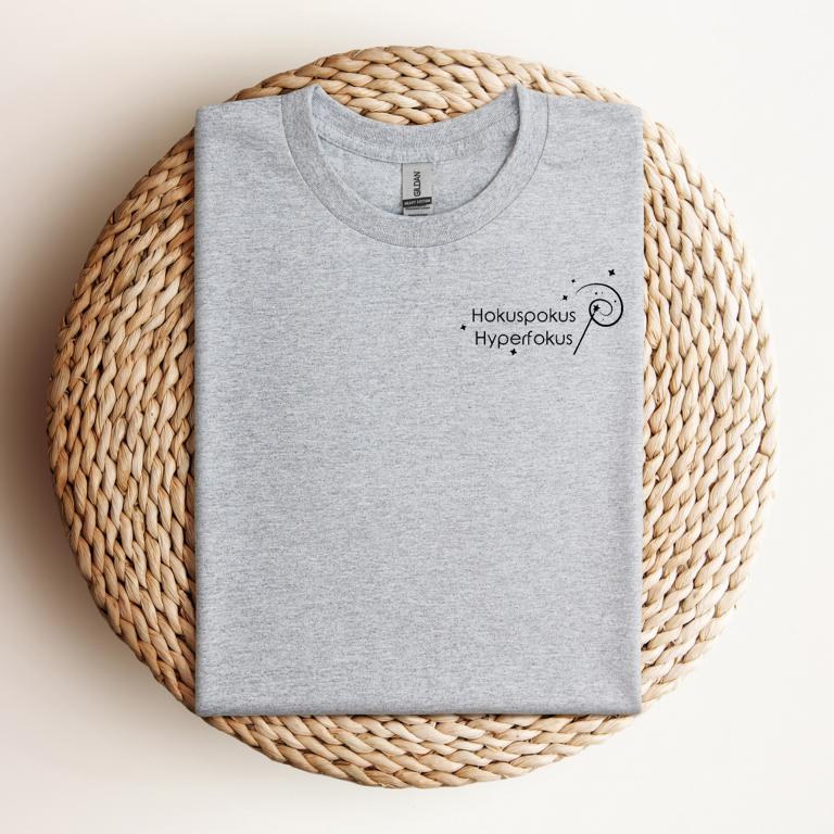 Gefaltetes T-Shirt mit minimalistischer Zauberstab-Grafik und dem Schriftzug „Hokuspokus Hyperfokus“ – humorvolles Design für ADHS & Neurodivergenz.